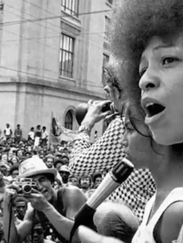 Angela Davis speaking to a large crowd during a civil rights rally, passionately addressing issues related to racial justice and systemic oppression.