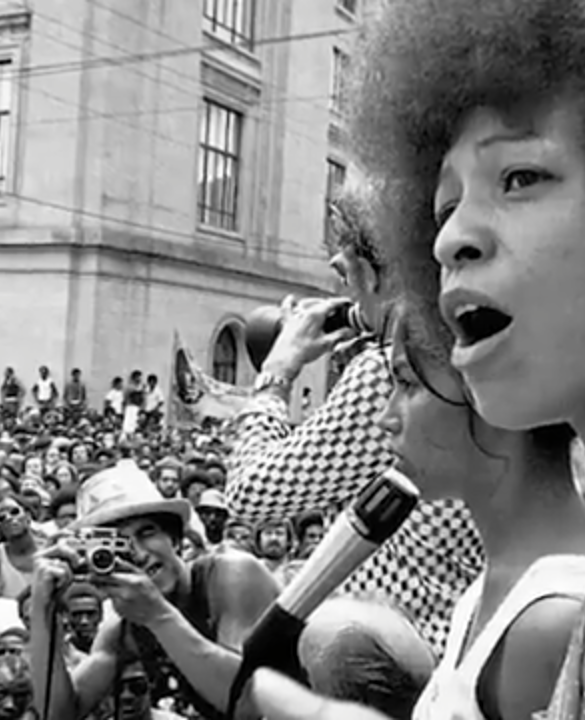 Angela Davis speaking to a large crowd during a civil rights rally, passionately addressing issues related to racial justice and systemic oppression.