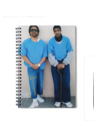 A collage of products featuring the image of prison artists C-Note and Joedee. The products include a phone case, spiral notebook, long-sleeve shirt, throw pillow, and tote bag. Each item displays the same photograph of the two artists standing side by side in blue prison uniforms, with C-Note on the left and Joedee on the right. The background of each product is a neutral gray, allowing the image to stand out prominently. These items are part of a merchandise collection available on BlackAugust.art.