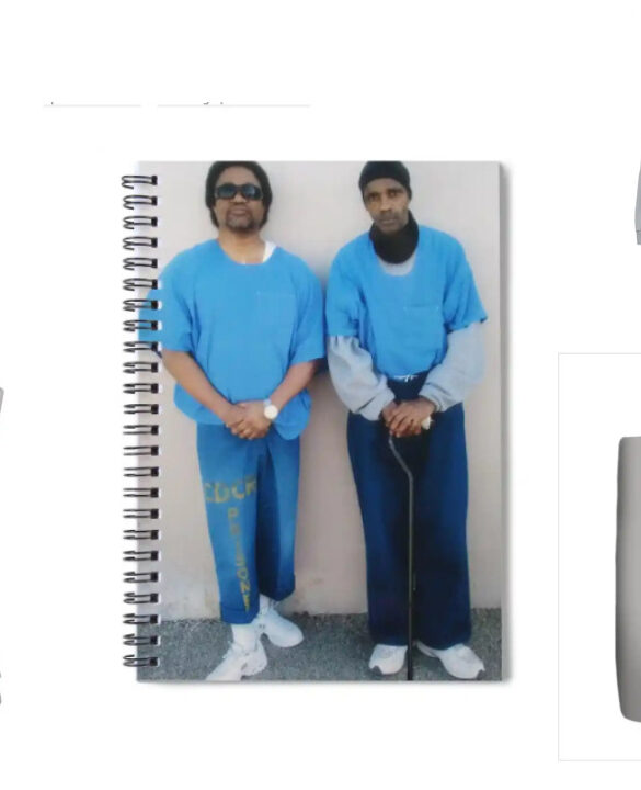 A collage of products featuring the image of prison artists C-Note and Joedee. The products include a phone case, spiral notebook, long-sleeve shirt, throw pillow, and tote bag. Each item displays the same photograph of the two artists standing side by side in blue prison uniforms, with C-Note on the left and Joedee on the right. The background of each product is a neutral gray, allowing the image to stand out prominently. These items are part of a merchandise collection available on BlackAugust.art.